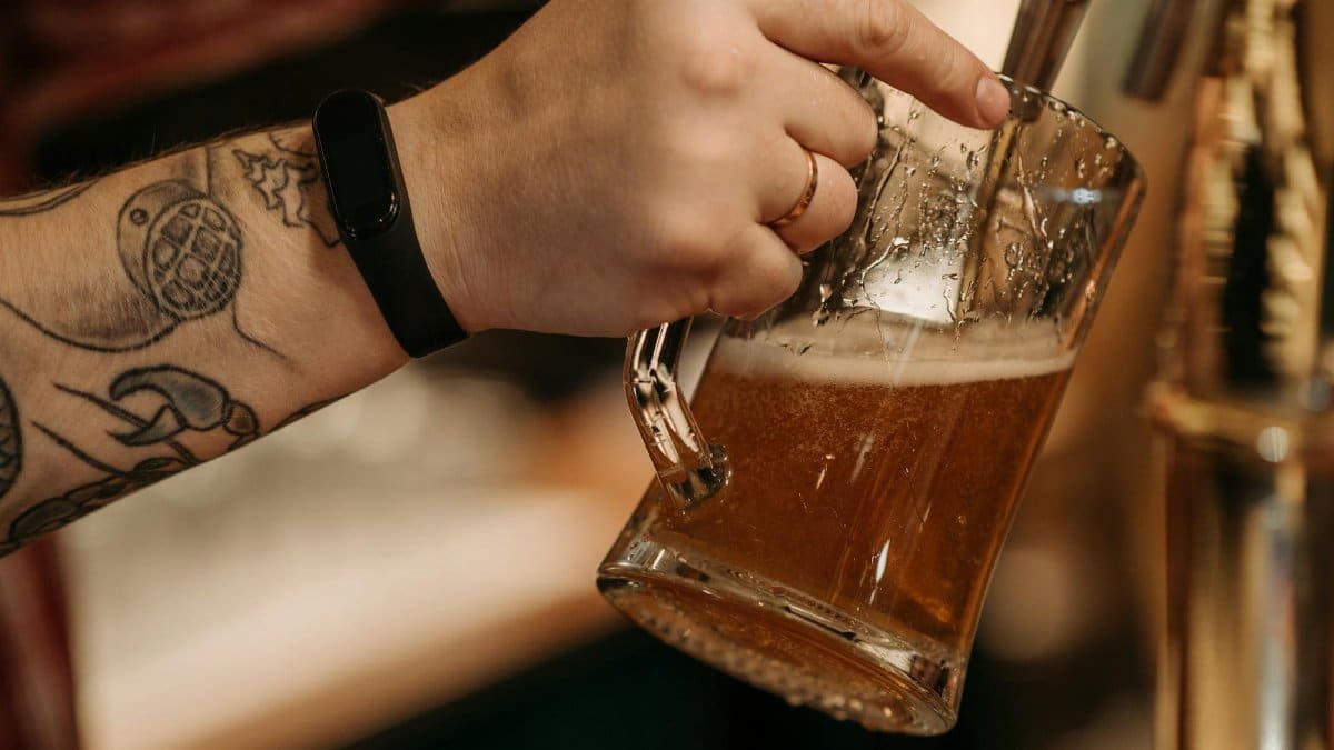 Close-up of a tattooed hand pouring beer into a glass mug from a draft tap.