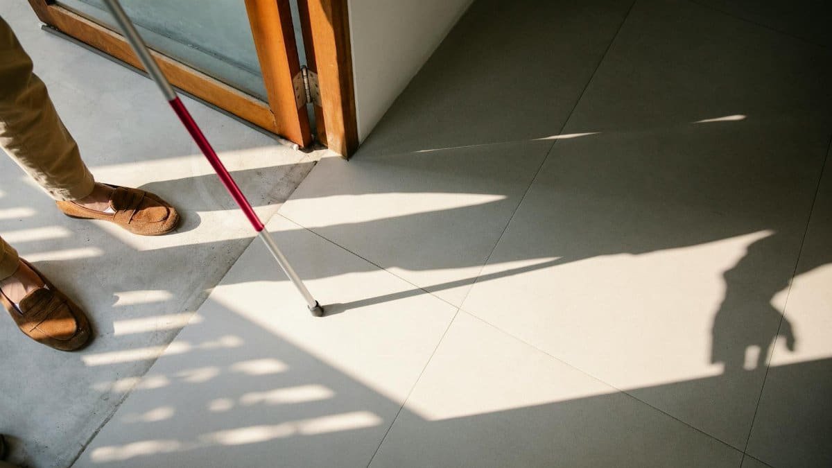 A person with a walking stick entering a sunlit room, casting shadows on tiled floor.