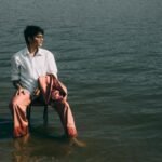 A man wearing a white shirt and pink suit pants sits in the water near a lake, exuding a sense of solitude and style.