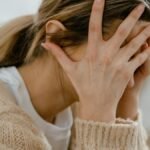 Woman sitting indoors with face covered by hands, expressing stress and frustration.