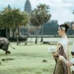 Elegant Cambodian couple in traditional attire at Angkor Wat with an elephant in the background.