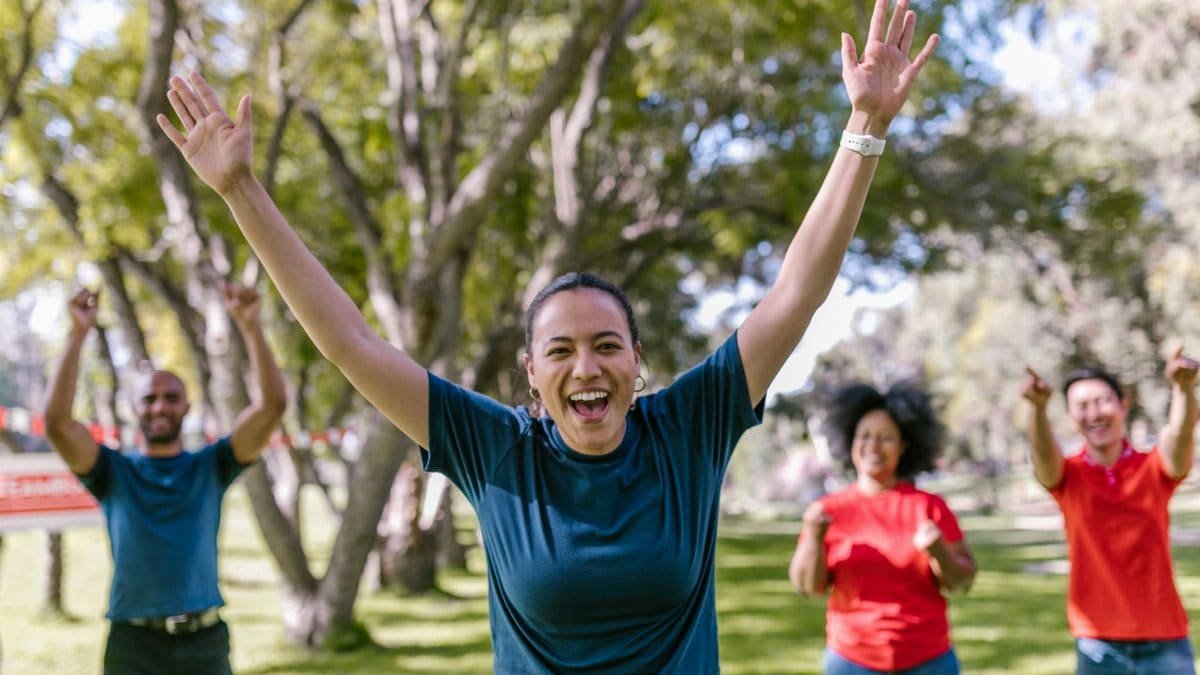 Diverse group of people celebrating success outdoors, with joyful expressions and raised arms in a park setting.