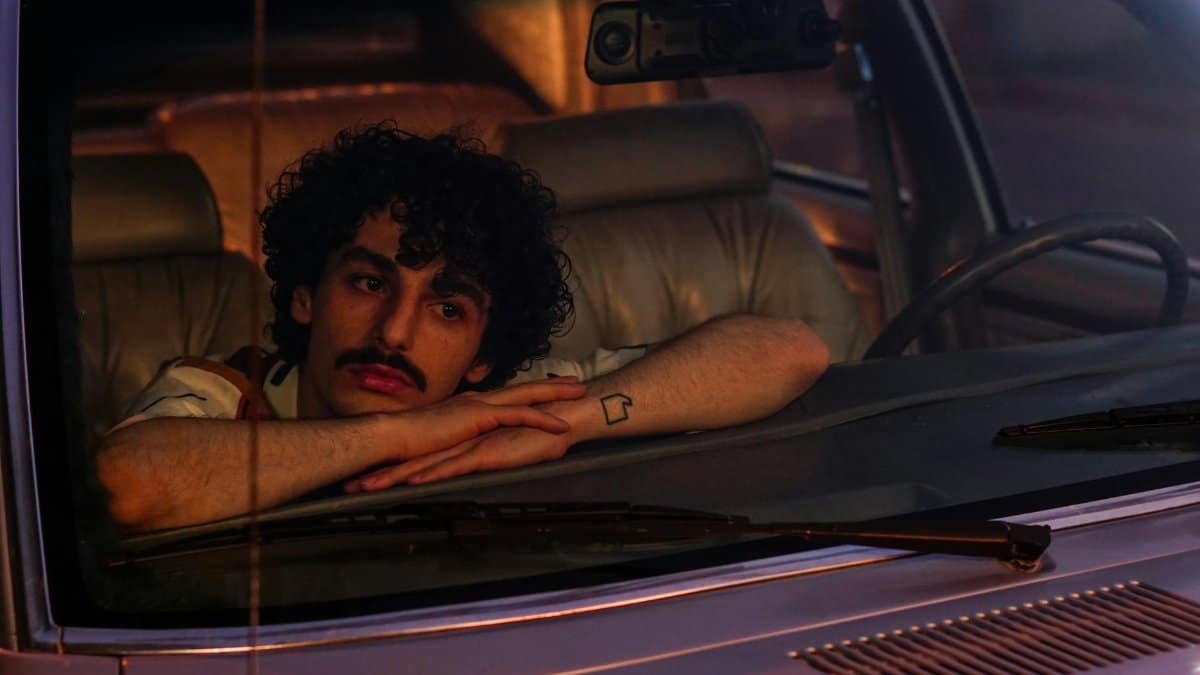 A man with curly hair and mustache, leaning in a car's interior, gazing thoughtfully at twilight.