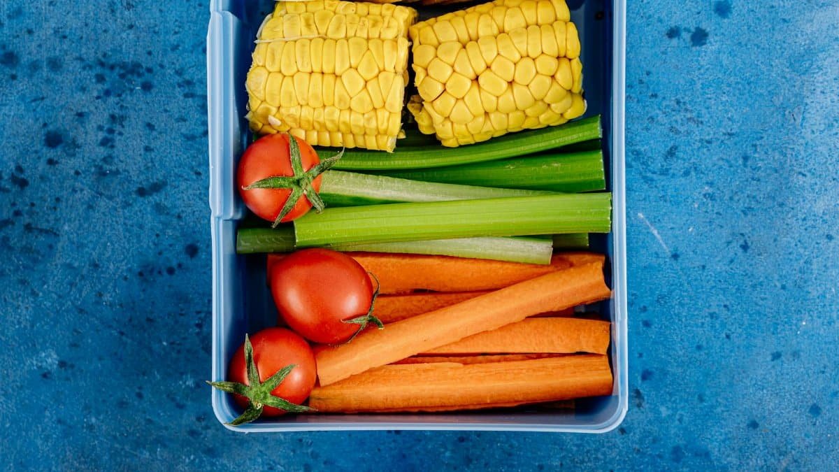 Top view of a lunchbox filled with vegetables, corn, tomatoes, and snacks on a blue background.