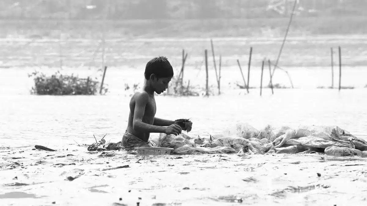 A young boy wades in the water, engaging in fishing, set in a tranquil and monochromatic landscape.