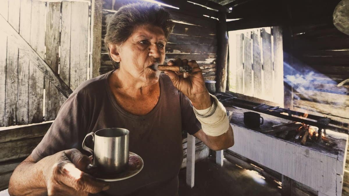 Elderly woman in a wooden house, enjoying a cigar with a tin cup indoors.