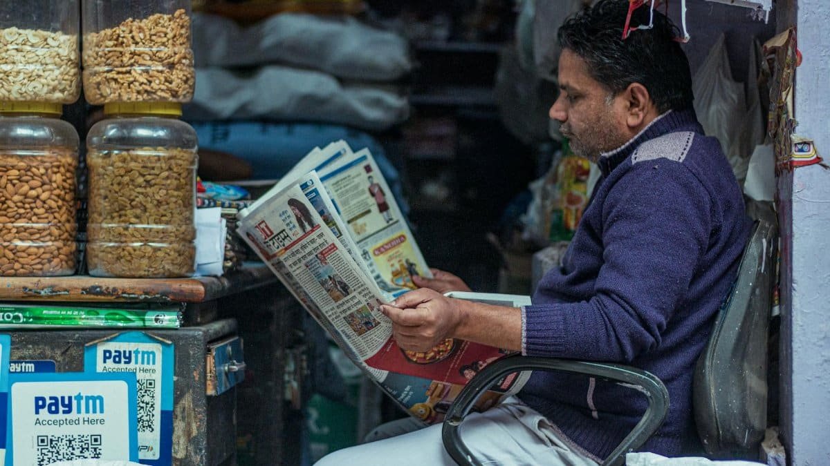 Man reading a newspaper in a small shop, surrounded by jars of snacks. Indoor market scene.