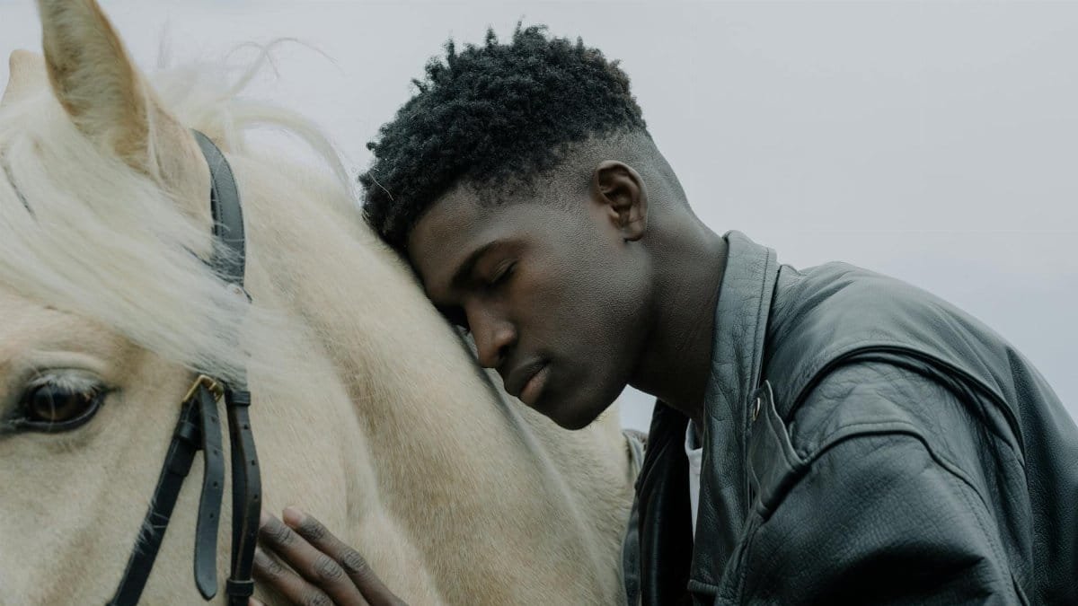 A young man rests his head affectionately on a white horse outdoors.