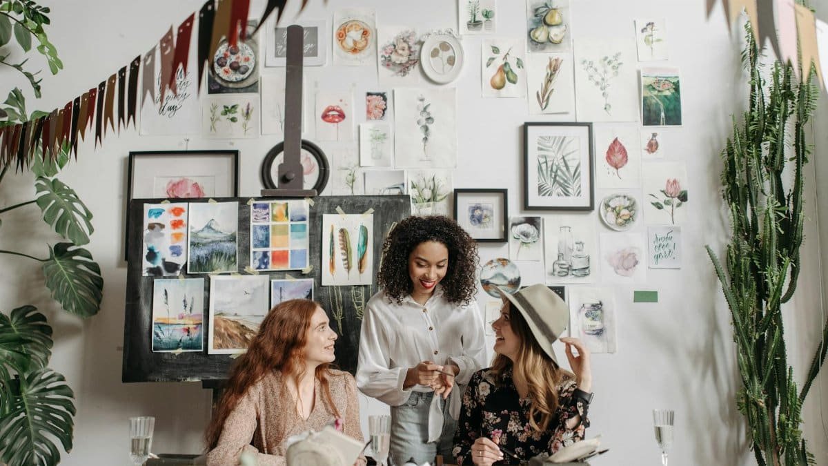 Three women enjoying a creative art workshop with paintings and plants around them.