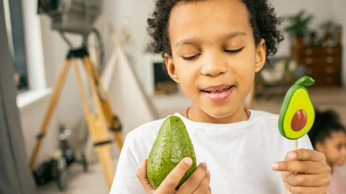 Smiling child holding an avocado and avocado-shaped lollipop in a cozy room.