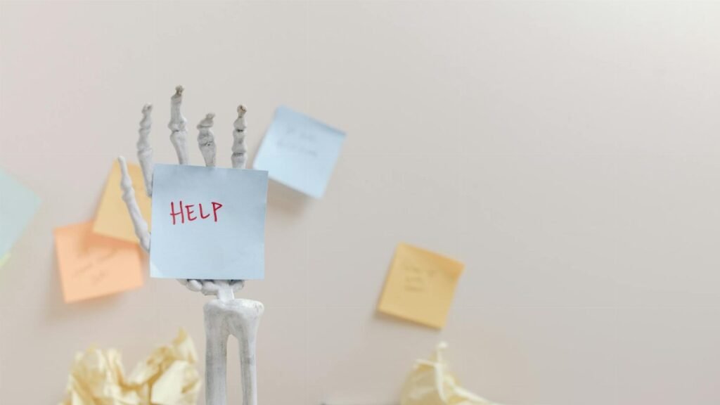 Skeleton hand holding a sticky note with 'HELP' written, against colorful notes background.
