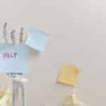 Skeleton hand holding a sticky note with 'HELP' written, against colorful notes background.