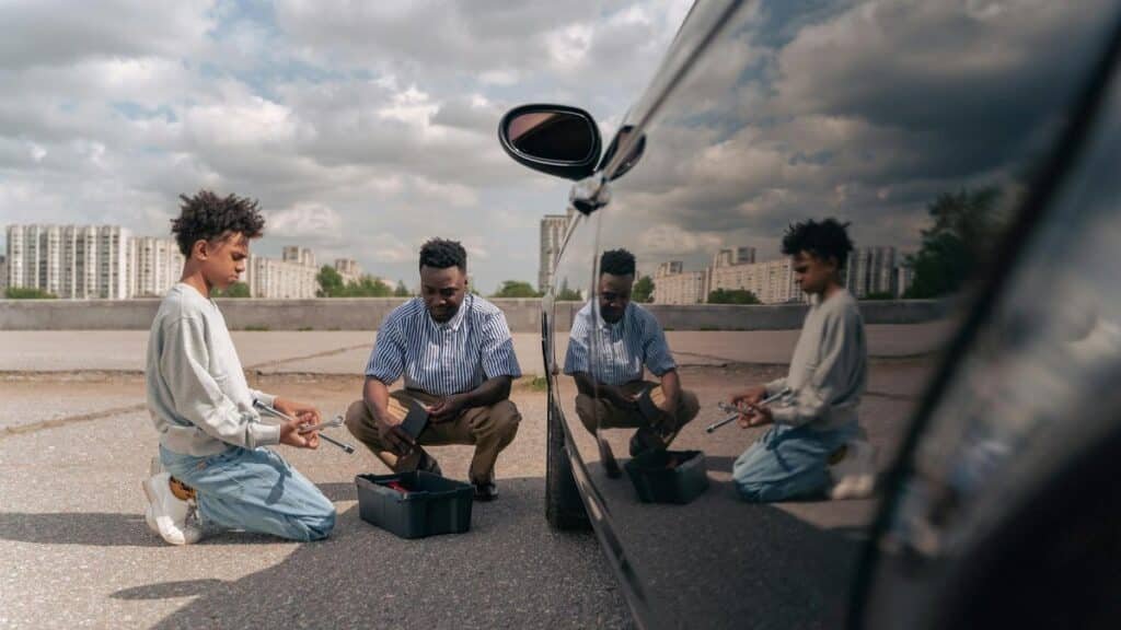 Father and son kneeling together outdoors, learning car maintenance with tools.