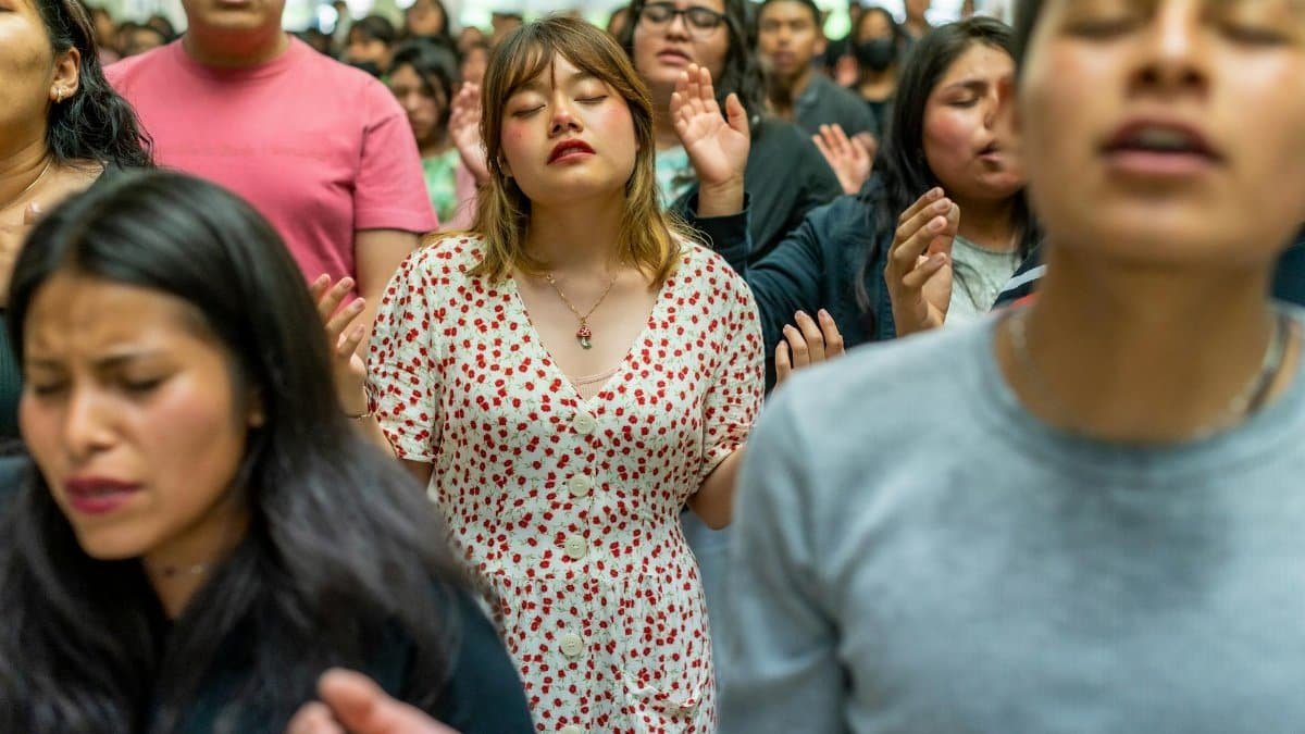A diverse group of people with eyes closed and hands raised participate in a heartfelt communal prayer indoors.