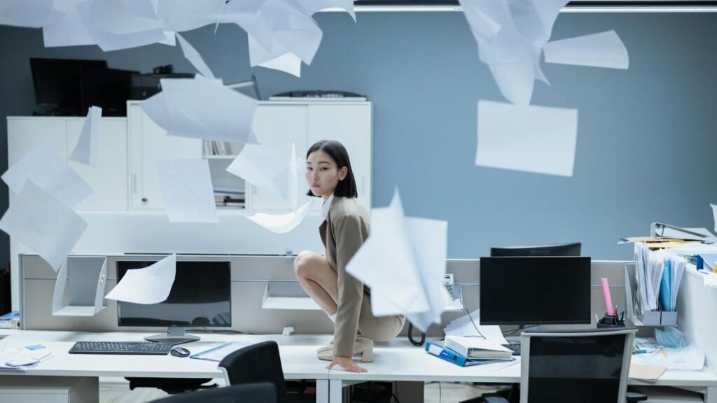 Woman perched on a desk in a chaotic office with papers flying, representing workplace stress.