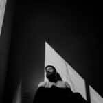 A bearded man sits indoors in dramatic lighting with shadows casting patterns.