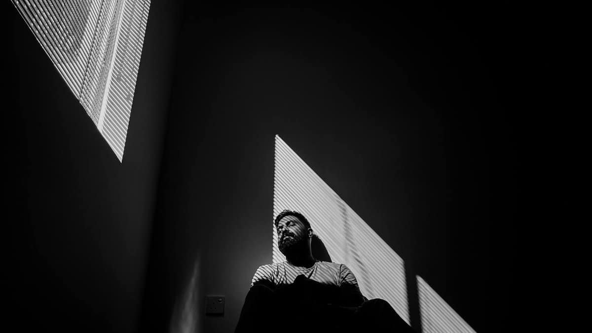 A bearded man sits indoors in dramatic lighting with shadows casting patterns.