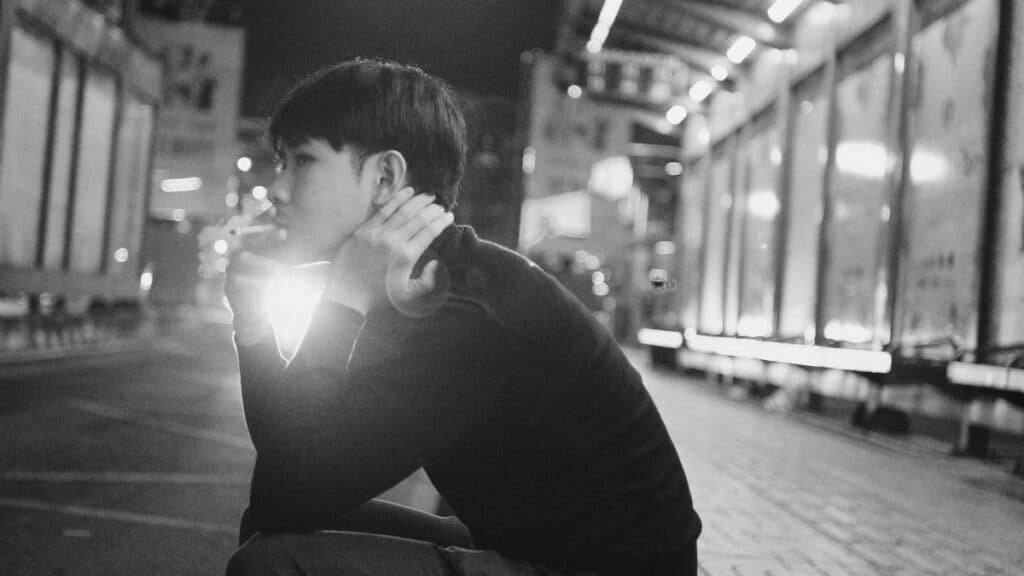 Black and white young ethnic man sitting on pavement and smoking cigarette in evening in city