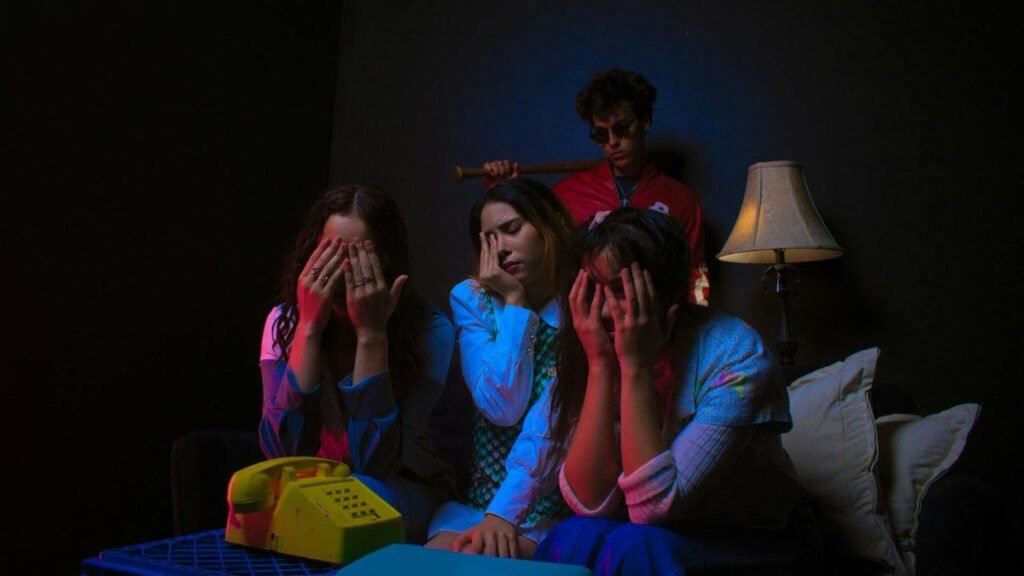 Three people sit with faces in hands indoors, conveying stress, while a man stands behind them.