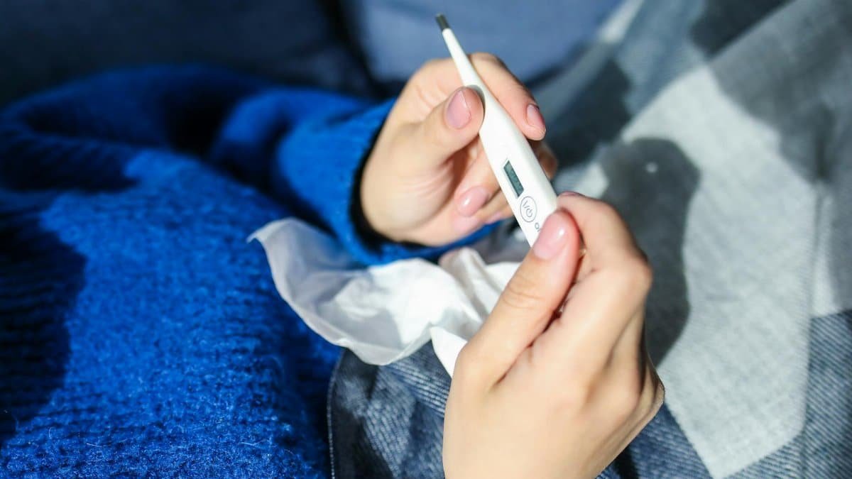 A woman checks a digital thermometer while lying indoors, showing signs of illness.