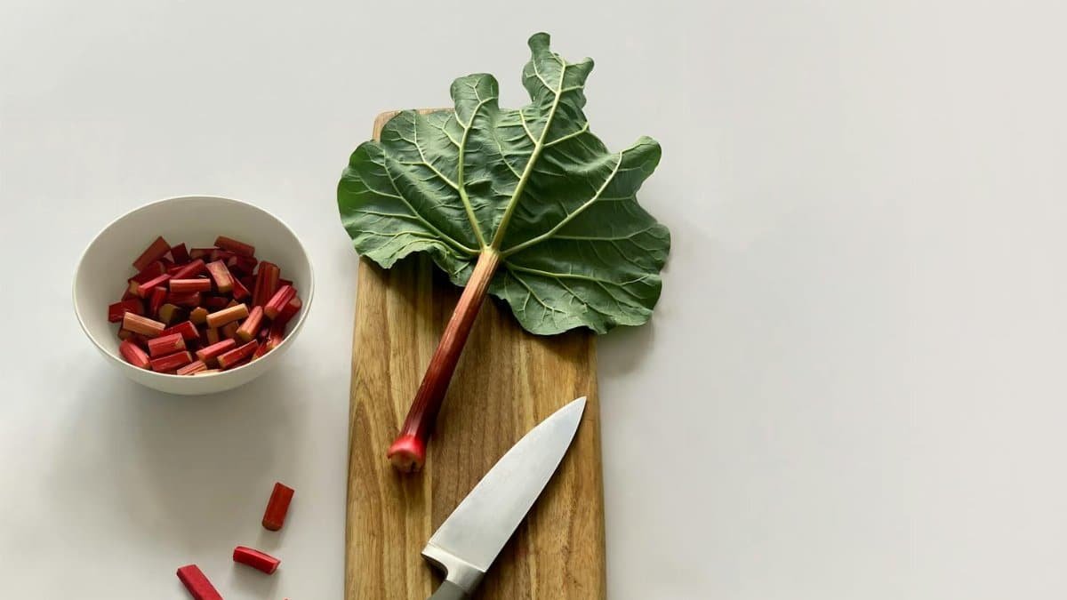 Fresh rhubarb stalk and leaves on a wooden cutting board with a knife, ready for cooking.