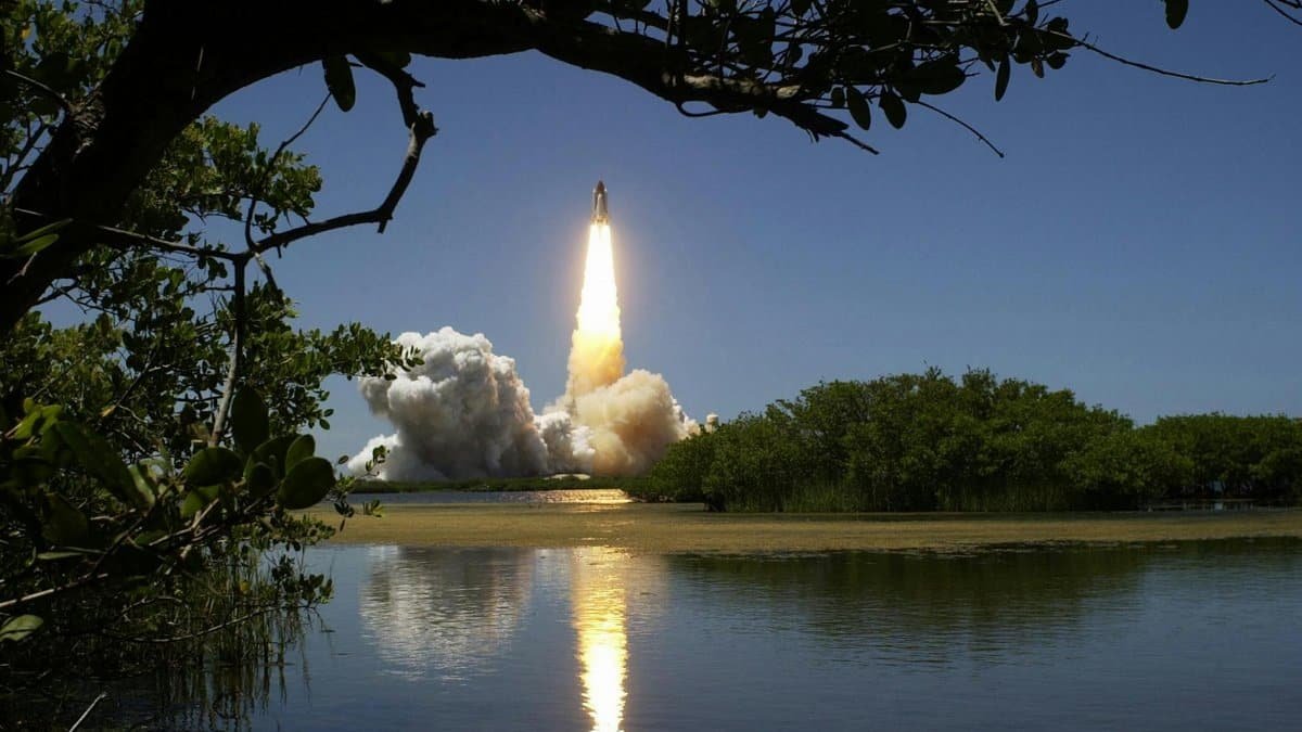 A dramatic rocket launch with smoke and flames, reflected in calm waters and framed by trees.