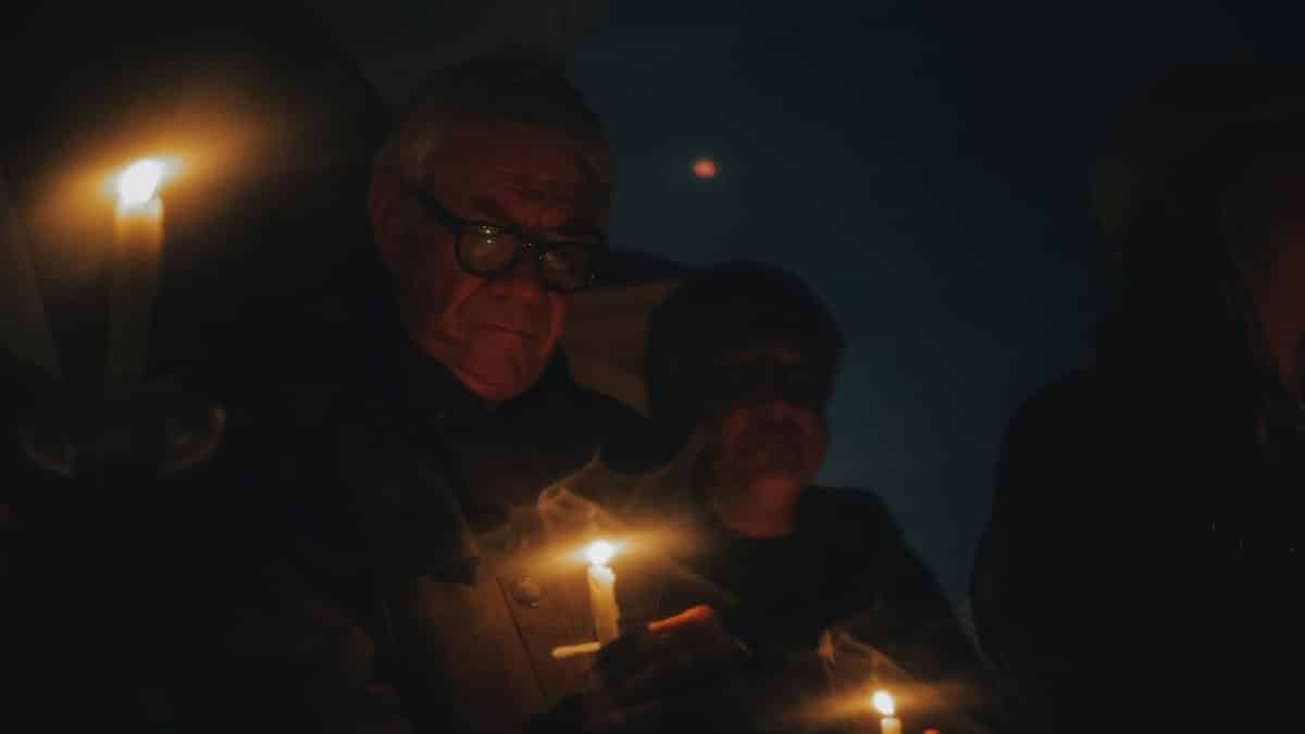 An elderly man holds a candle during a nighttime vigil, creating a moody and introspective atmosphere.