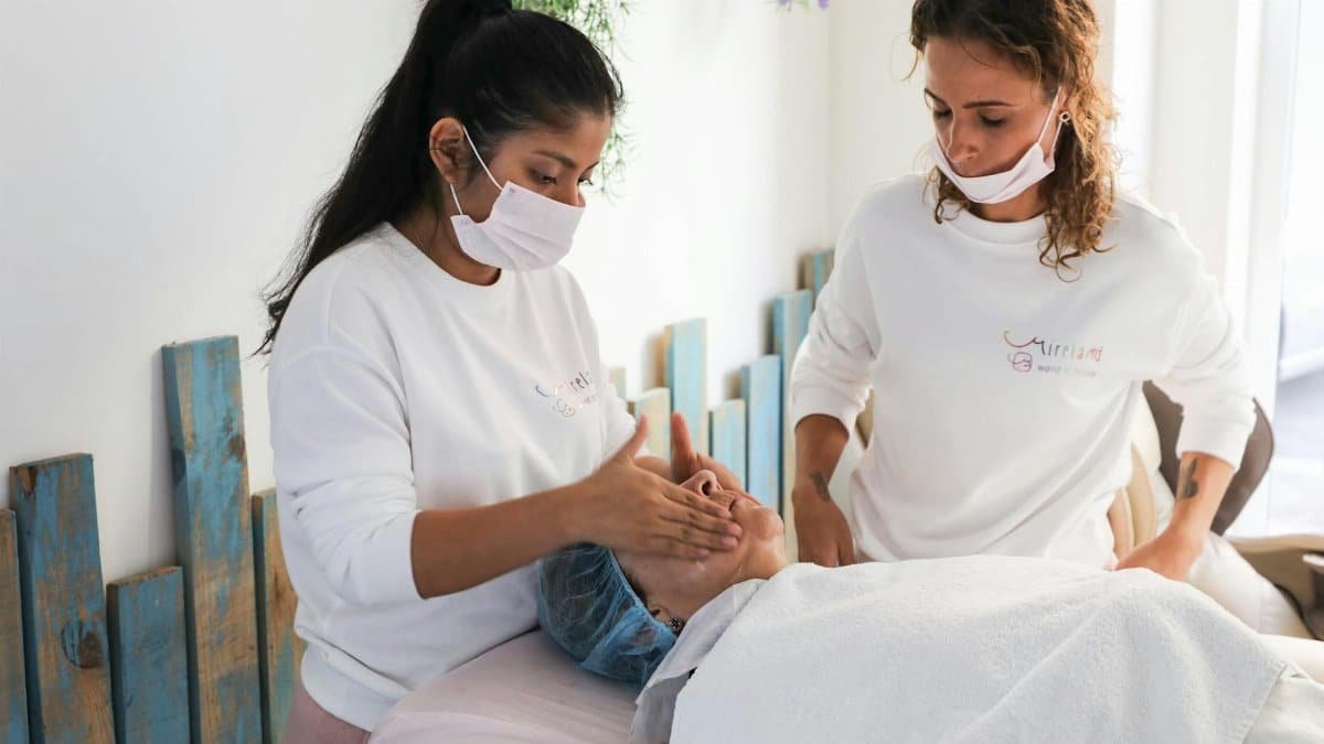 Two women in a relaxing spa setting conducting a facial massage on a client. Indoors, calm atmosphere.