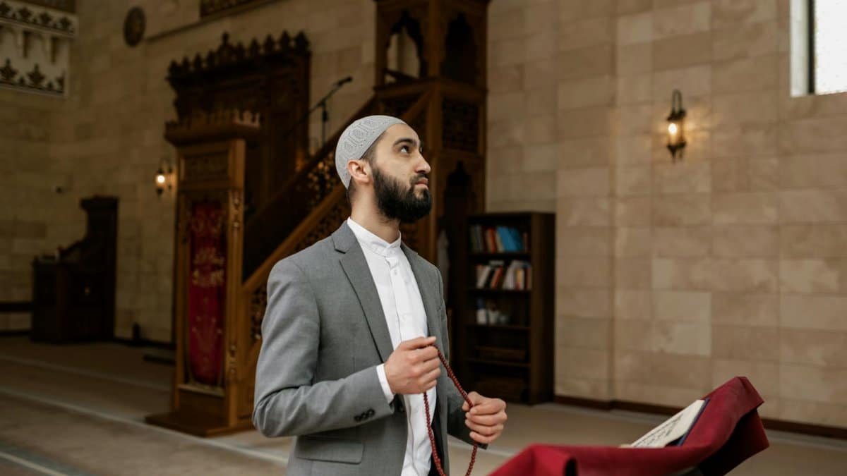 Man wearing a suit and skullcap holds prayer beads in an ornate mosque interior during Ramadan.