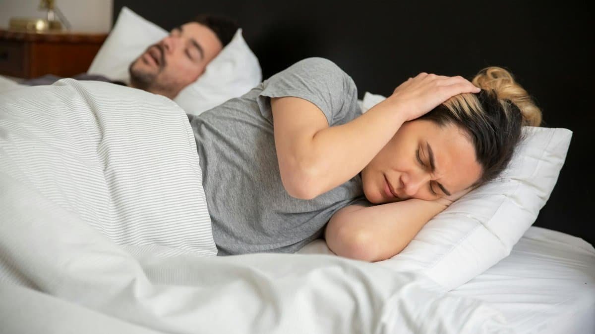 A woman looks frustrated in bed as her partner snores loudly, highlighting sleep disturbance issues.