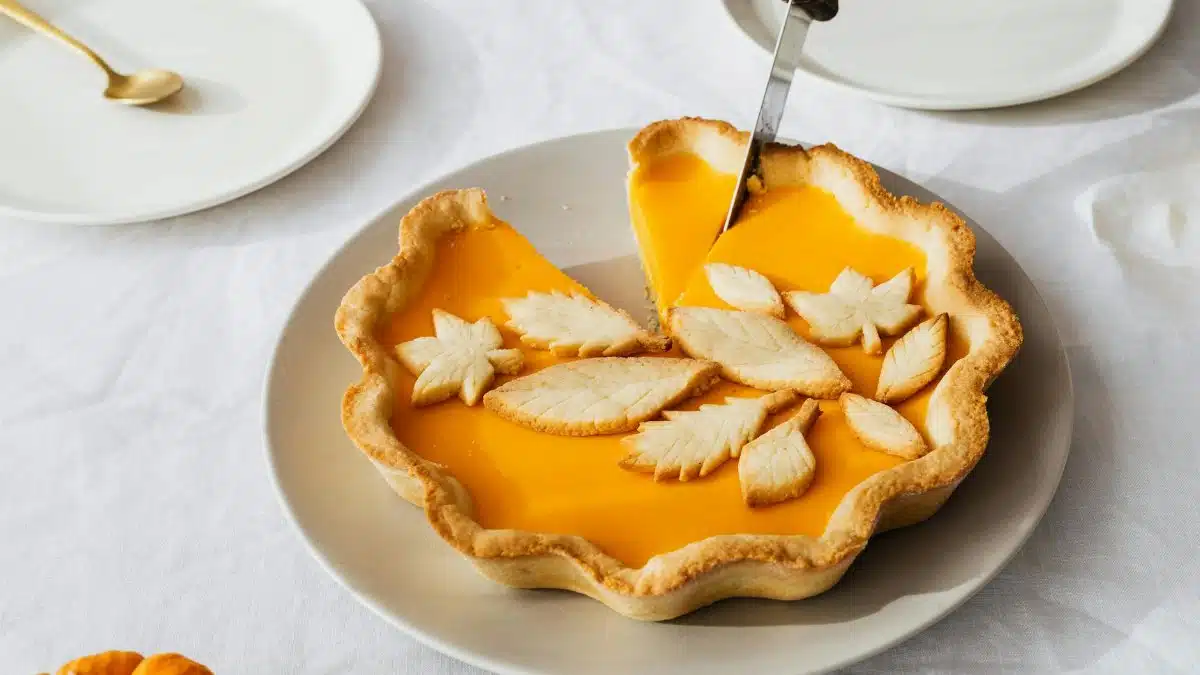 A beautiful pumpkin pie with leaf decorations being sliced on a dining table.