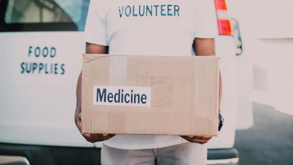 A volunteer holding a box labeled medicine near a delivery van marked food supplies.