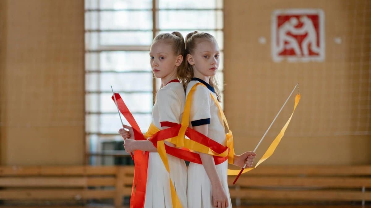 Young twin girls practicing rhythmic gymnastics with ribbons inside a sport hall.