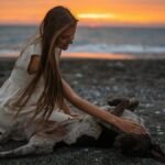 Young woman in white dress petting a dog on a beach at sunset, capturing a serene moment. Alakhadzi, Georgia.