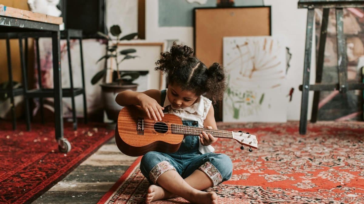 Adorable young girl sitting on the floor playing a ukulele in a vibrant art studio.