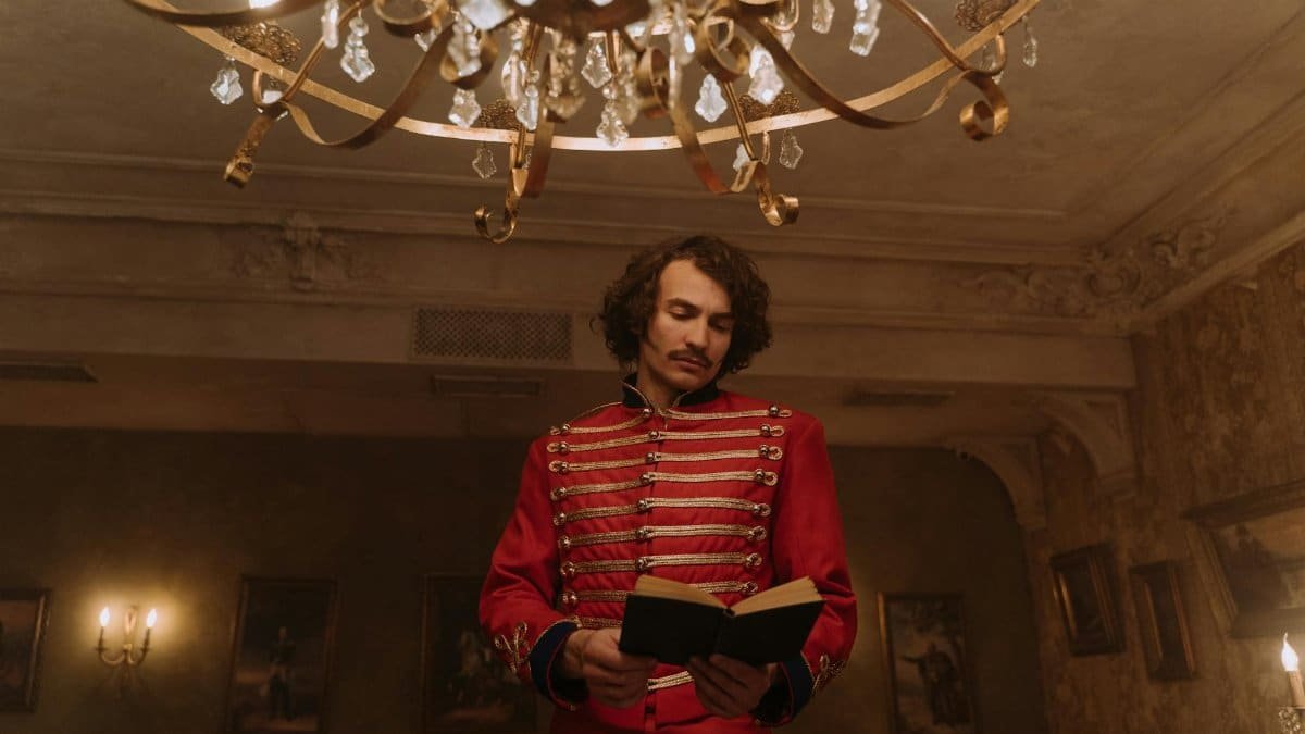 A man in a historic red coat reads a book under a chandelier indoors.