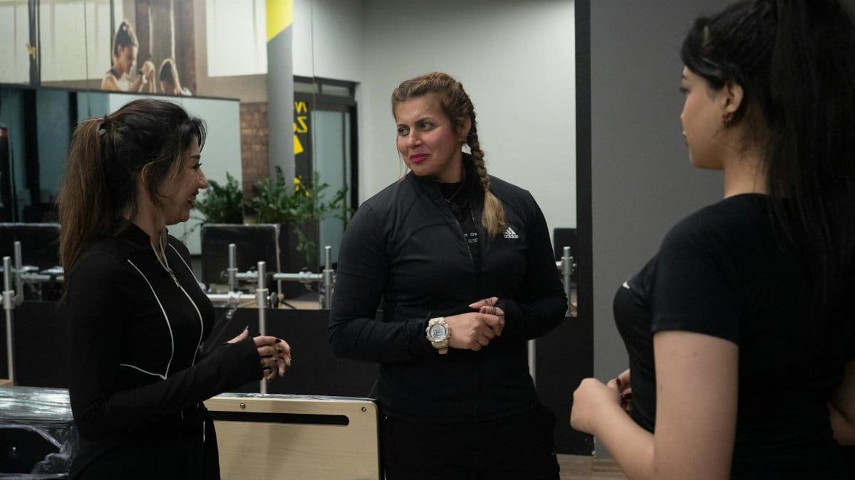 Three women in gym attire discuss fitness inside a modern health club.