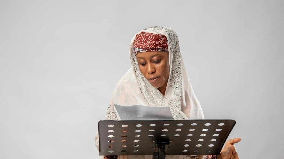 An African woman in traditional clothing reads from a music stand, expressing focus and grace.