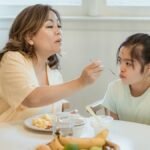 A mother lovingly feeds her daughter breakfast in a cozy home setting.