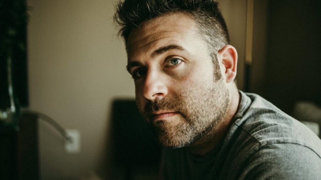 Close-up portrait of a thoughtful bearded man indoors with moody lighting.