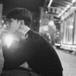 Black and white young ethnic man sitting on pavement and smoking cigarette in evening in city