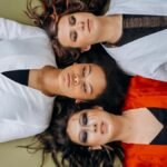 A peaceful scene of three women lying down with eyes closed, wearing colorful clothing.