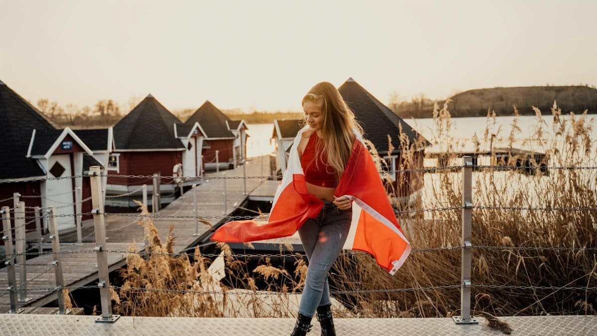 Caucasian woman smiling by lakeside cabins in Toronto, holding a Canadian flag at sunset.