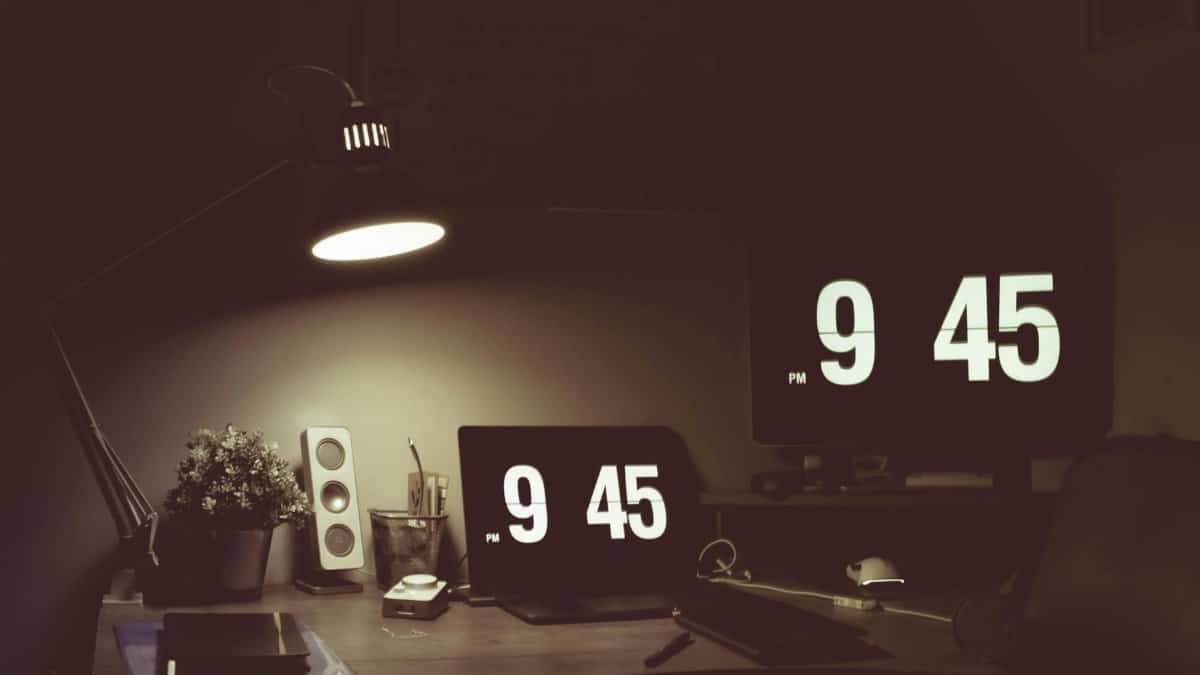 A cozy desk setup featuring digital clocks, a desk lamp, and modern devices in low light.