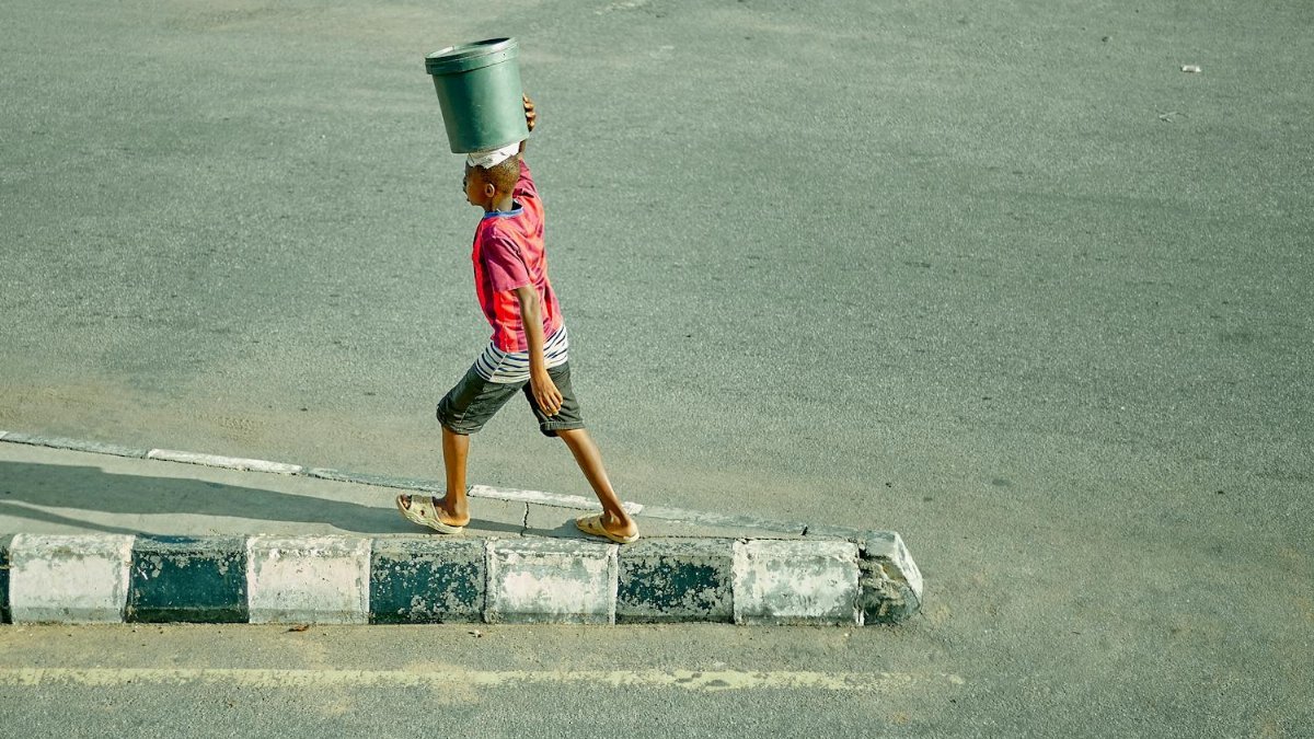 A child in a pink shirt carries a bucket on head while walking on urban pavement.
