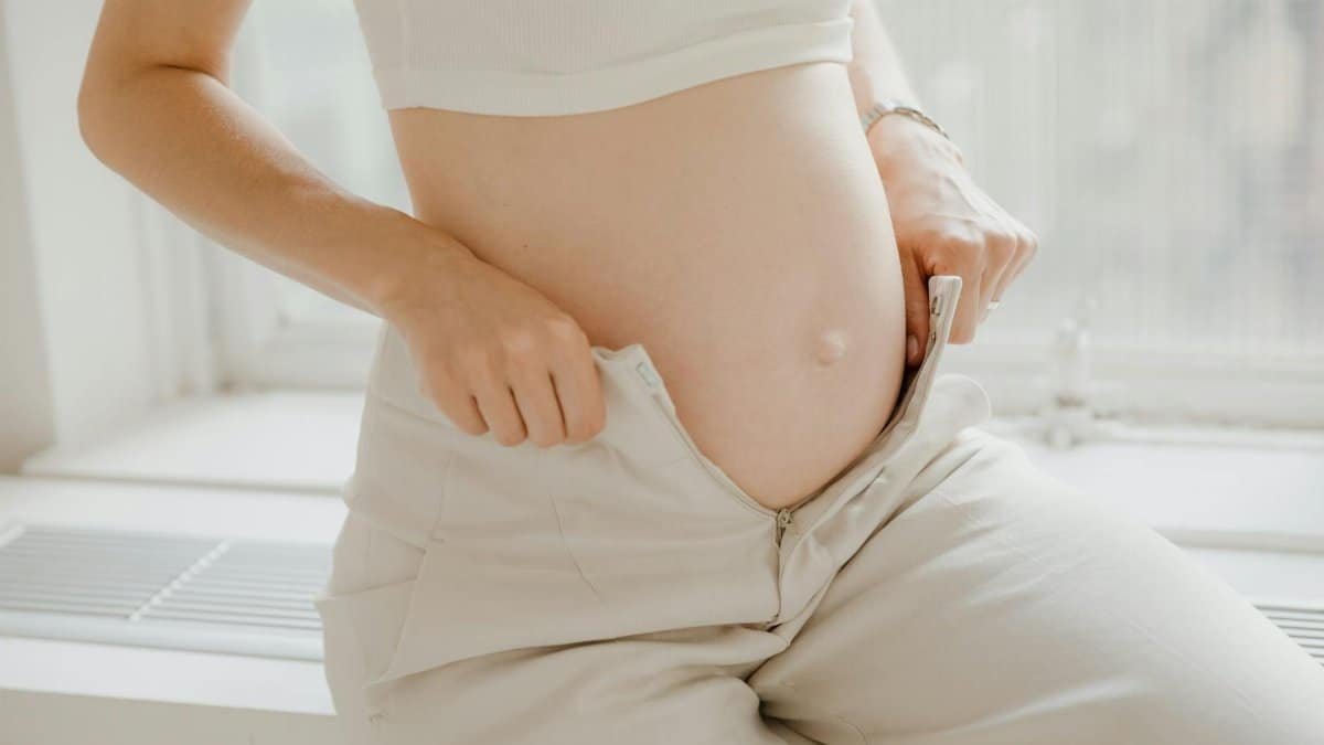 A close-up of a pregnant woman adjusting her pants, highlighting her belly in a sunlit room.