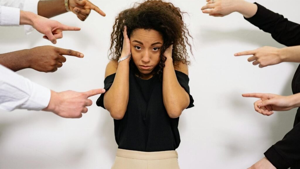 Stressed woman with hands on ears surrounded by pointing fingers, illustrating pressure.