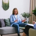 Worried male patient sitting on couch while having conversation about problems with unrecognizable professional psychologist during psychotherapy appointment in office