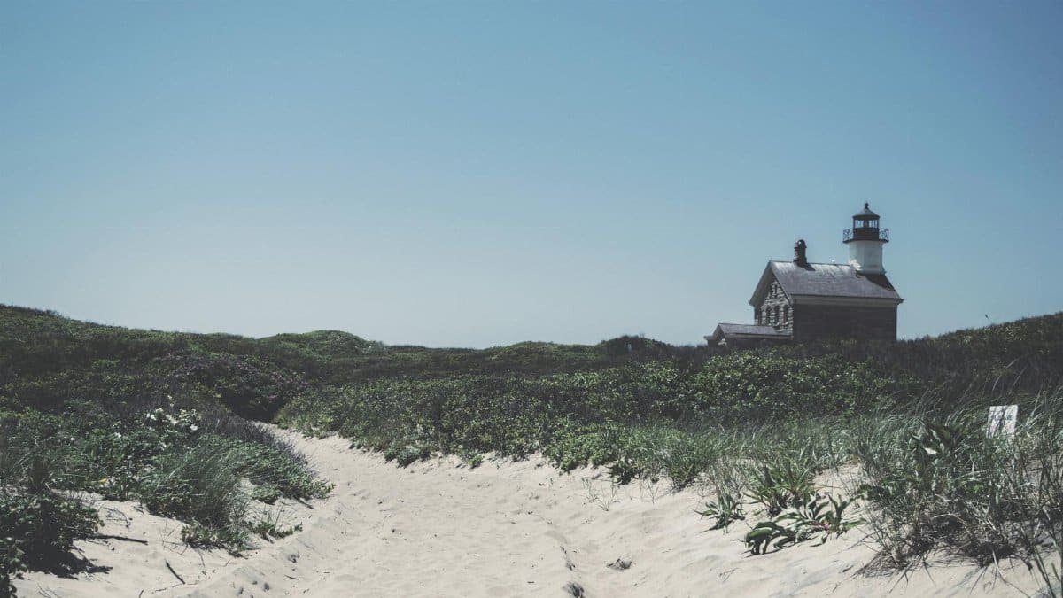 Scenic view of Block Island Lighthouse with sandy path and lush green dunes under blue sky.