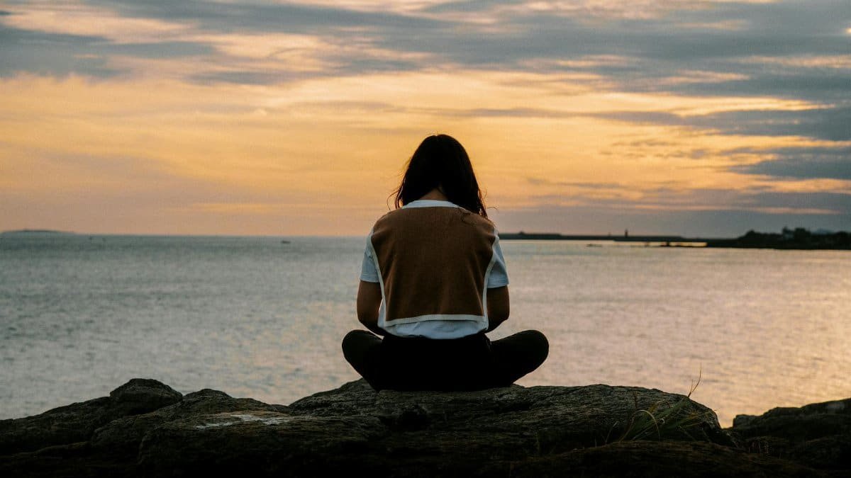 A serene scene of a woman sitting on rocks by the sea during a vibrant sunset.